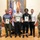 A formal military awards ceremony featuring seven individuals posing for a group photograph in what appears to be an official government or military building. Three civilians in the center are holding framed award certificates, flanked by military personnel in Army Combat Uniforms (ACU/OCP) on both ends and two civilians. Flags including the U.S. flag are visible in the background along with a large framed painting.
