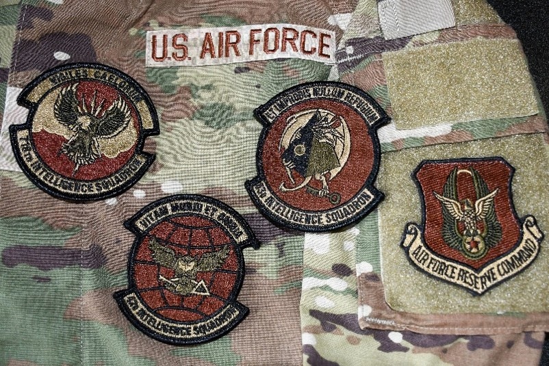 The image shows a U.S. Air Force OCP (Operational Camouflage Pattern) uniform with multiple embroidered patches displayed. Four circular intelligence squadron patches and one shield-shaped command patch are visible, along with a 'U.S. AIR FORCE' tape. The patches are associated with intelligence squadrons and Air Force Reserve Command.