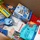 A cardboard box filled with a variety of packaged food and personal care items, likely assembled as a care package or donation box. The contents include snack foods, hydration products, and hygiene items packed loosely together. The assortment suggests this may be a gift box, emergency supply kit, or charitable donation package.