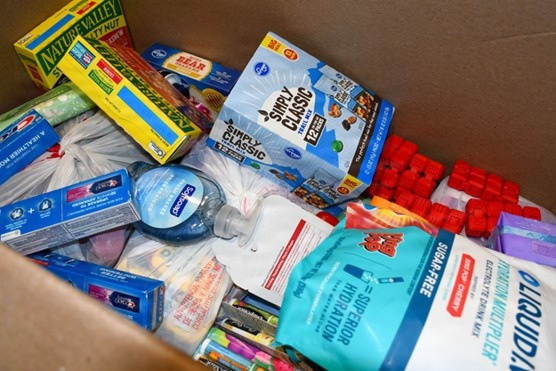 A cardboard box filled with a variety of packaged food and personal care items, likely assembled as a care package or donation box. The contents include snack foods, hydration products, and hygiene items packed loosely together. The assortment suggests this may be a gift box, emergency supply kit, or charitable donation package.