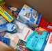 A cardboard box filled with a variety of packaged food and personal care items, likely assembled as a care package or donation box. The contents include snack foods, hydration products, and hygiene items packed loosely together. The assortment suggests this may be a gift box, emergency supply kit, or charitable donation package.
