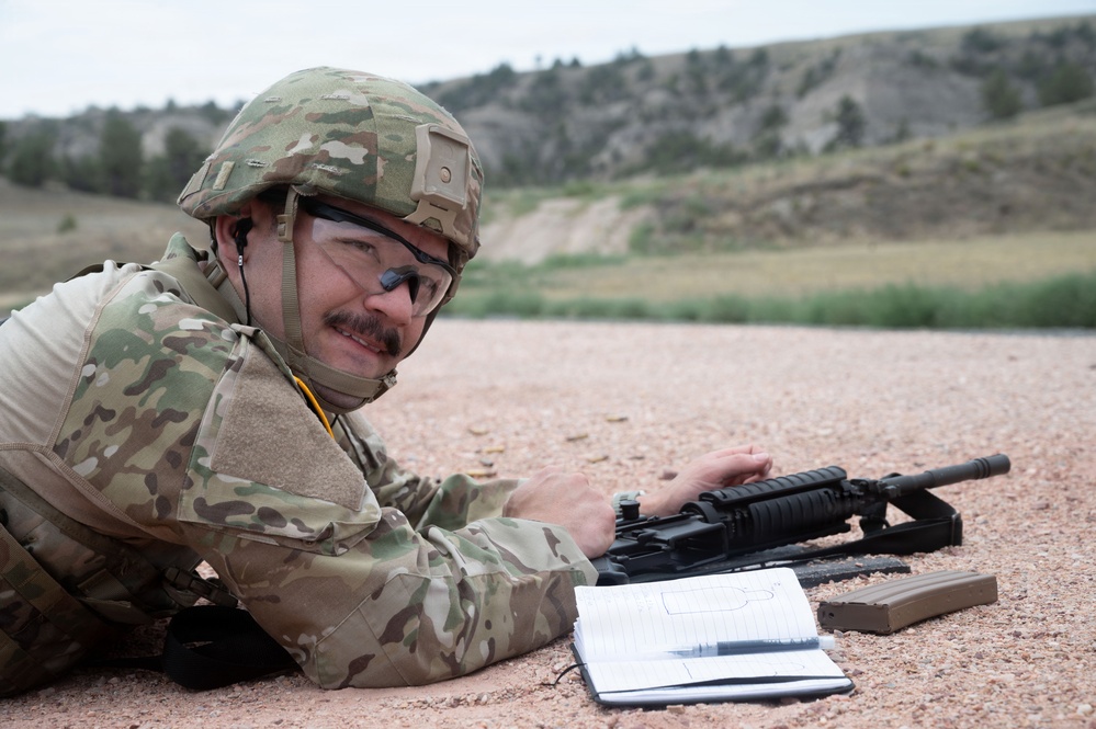 A soldier in MultiCam/OCP camouflage uniform is lying prone on a gravel shooting range, smiling toward the camera. They are equipped with a combat helmet, ballistic eyewear, and body armor, with an M4/M16-style rifle and a detached magazine in front of them. A notepad with what appears to be a target scoring sheet is also visible on the ground beside the weapon.