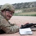 A soldier in MultiCam/OCP camouflage uniform is lying prone on a gravel shooting range, smiling toward the camera. They are equipped with a combat helmet, ballistic eyewear, and body armor, with an M4/M16-style rifle and a detached magazine in front of them. A notepad with what appears to be a target scoring sheet is also visible on the ground beside the weapon.