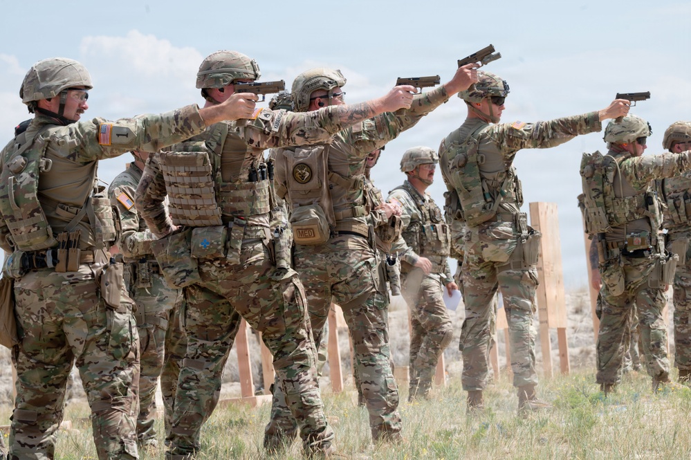 A group of military personnel in MultiCam/OCP camouflage uniforms are conducting pistol qualification or training at an outdoor firing range. Multiple soldiers are simultaneously aiming semi-automatic pistols downrange in an extended shooting stance. The setting appears to be an open, arid training range with wooden target stands visible in the background.