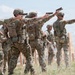 A group of military personnel in MultiCam/OCP camouflage uniforms are conducting pistol qualification or training at an outdoor firing range. Multiple soldiers are simultaneously aiming semi-automatic pistols downrange in an extended shooting stance. The setting appears to be an open, arid training range with wooden target stands visible in the background.