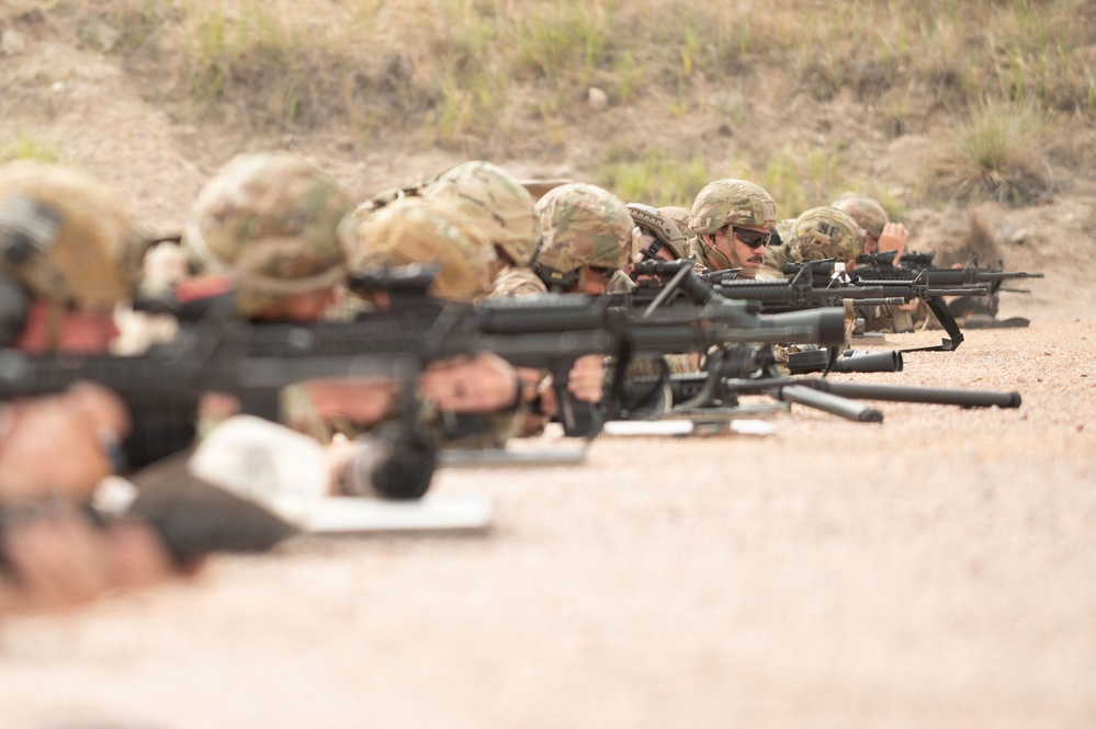A line of military personnel in full combat gear are in the prone firing position on a shooting range. They are equipped with rifles mounted on bipods and wearing camouflage uniforms and helmets. The image appears to depict a live-fire training exercise.