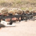 A line of military personnel in full combat gear are in the prone firing position on a shooting range. They are equipped with rifles mounted on bipods and wearing camouflage uniforms and helmets. The image appears to depict a live-fire training exercise.