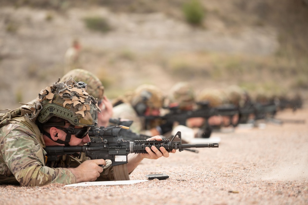 Multiple military personnel are in a prone firing position on what appears to be an outdoor shooting range. The foreground soldier is clearly visible in full combat gear, aiming an M4/M16-style carbine downrange. The image depicts a live-fire training exercise with several shooters aligned in a row extending into the background.