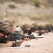 Multiple military personnel are in a prone firing position on what appears to be an outdoor shooting range. The foreground soldier is clearly visible in full combat gear, aiming an M4/M16-style carbine downrange. The image depicts a live-fire training exercise with several shooters aligned in a row extending into the background.