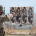 A U.S. Army soldier in full OCP (Operational Camouflage Pattern) uniform and helmet is firing a semi-automatic pistol at a military shooting range. The soldier is engaging multiple standard E-type silhouette paper targets mounted on wooden stands. A spent brass casing is visibly ejecting from the pistol, indicating the weapon has just been fired.