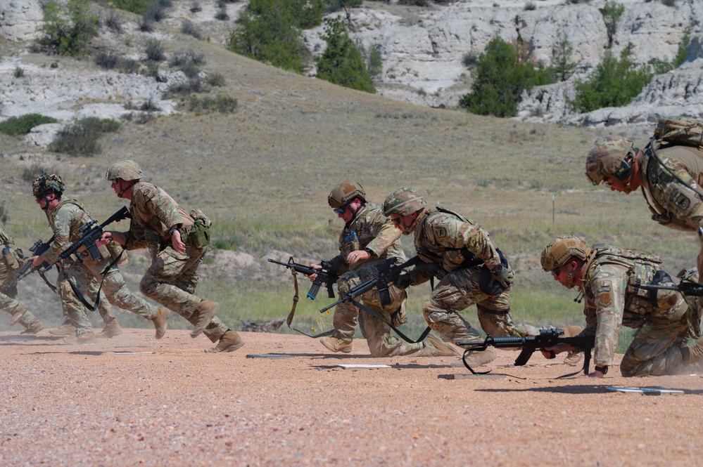 A group of military personnel in camouflage uniforms are conducting a tactical training exercise on a dirt range. The soldiers are moving in a low crouched formation while carrying rifles, suggesting a bounding or assault drill. The setting appears to be an arid training range with rocky bluffs and sparse vegetation in the background.