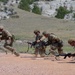 A group of military personnel in camouflage uniforms are conducting a tactical training exercise on a dirt range. The soldiers are moving in a low crouched formation while carrying rifles, suggesting a bounding or assault drill. The setting appears to be an arid training range with rocky bluffs and sparse vegetation in the background.