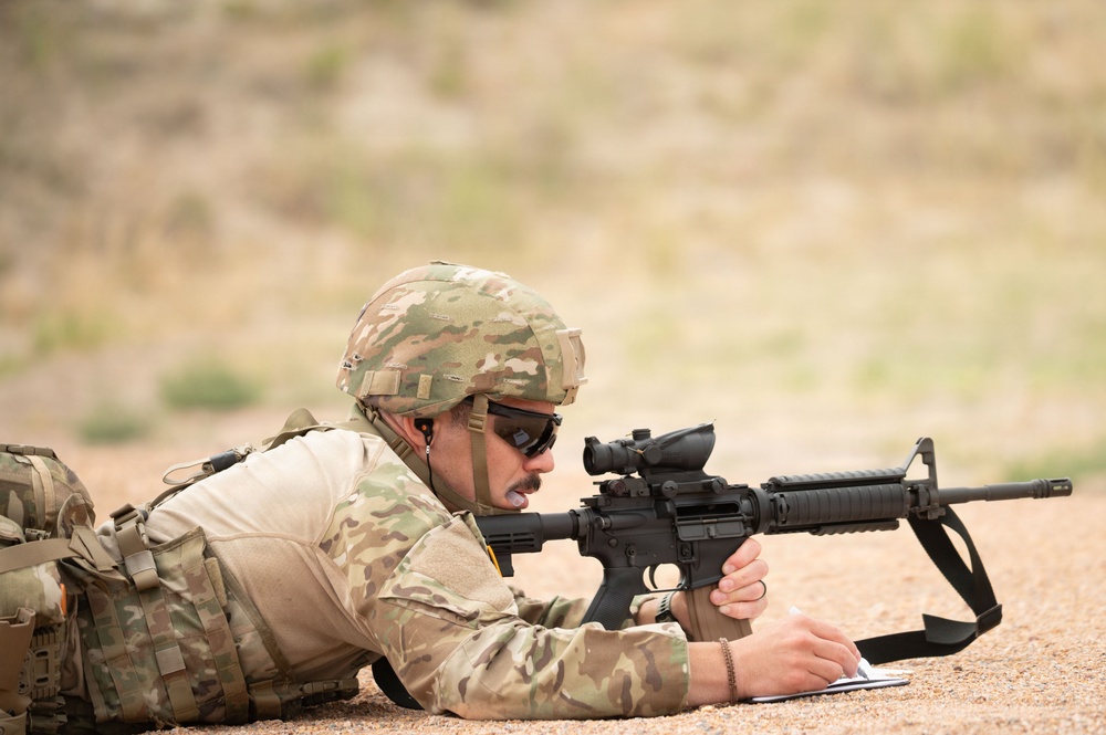 A soldier in full combat gear is lying prone on a dirt surface, aiming a rifle downrange while simultaneously writing on a notepad. The individual is wearing Operational Camouflage Pattern (OCP) uniform, a tactical helmet, ballistic sunglasses, and a plate carrier vest. This appears to be a training exercise or qualification range event.