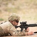 A soldier in full combat gear is lying prone on a dirt surface, aiming a rifle downrange while simultaneously writing on a notepad. The individual is wearing Operational Camouflage Pattern (OCP) uniform, a tactical helmet, ballistic sunglasses, and a plate carrier vest. This appears to be a training exercise or qualification range event.