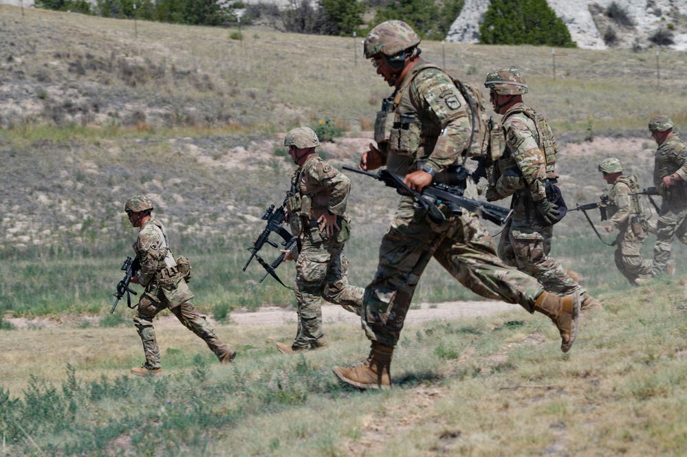 Multiple soldiers in OCP (Operational Camouflage Pattern) uniforms are running across an open grassy field during what appears to be a military training exercise. They are carrying assault rifles and wearing full combat gear including helmets, body armor, and tactical vests. The terrain features sparse vegetation, rocky outcroppings, and rolling hills in the background.