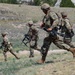 Multiple soldiers in OCP (Operational Camouflage Pattern) uniforms are running across an open grassy field during what appears to be a military training exercise. They are carrying assault rifles and wearing full combat gear including helmets, body armor, and tactical vests. The terrain features sparse vegetation, rocky outcroppings, and rolling hills in the background.