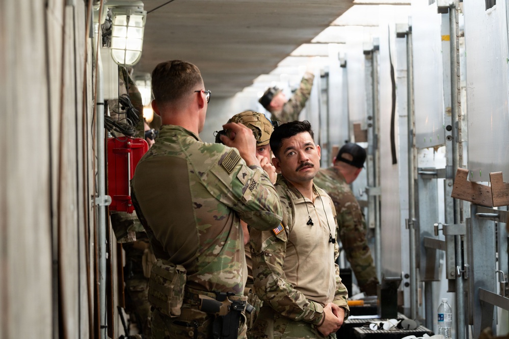 Military personnel in camouflage uniforms are working in a narrow corridor between what appear to be shipping containers or military equipment bays. One soldier appears to be assisting another with headgear or equipment adjustment, while additional personnel work in the background. The setting suggests a logistics, loading, or equipment preparation operation.
