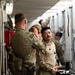 Military personnel in camouflage uniforms are working in a narrow corridor between what appear to be shipping containers or military equipment bays. One soldier appears to be assisting another with headgear or equipment adjustment, while additional personnel work in the background. The setting suggests a logistics, loading, or equipment preparation operation.