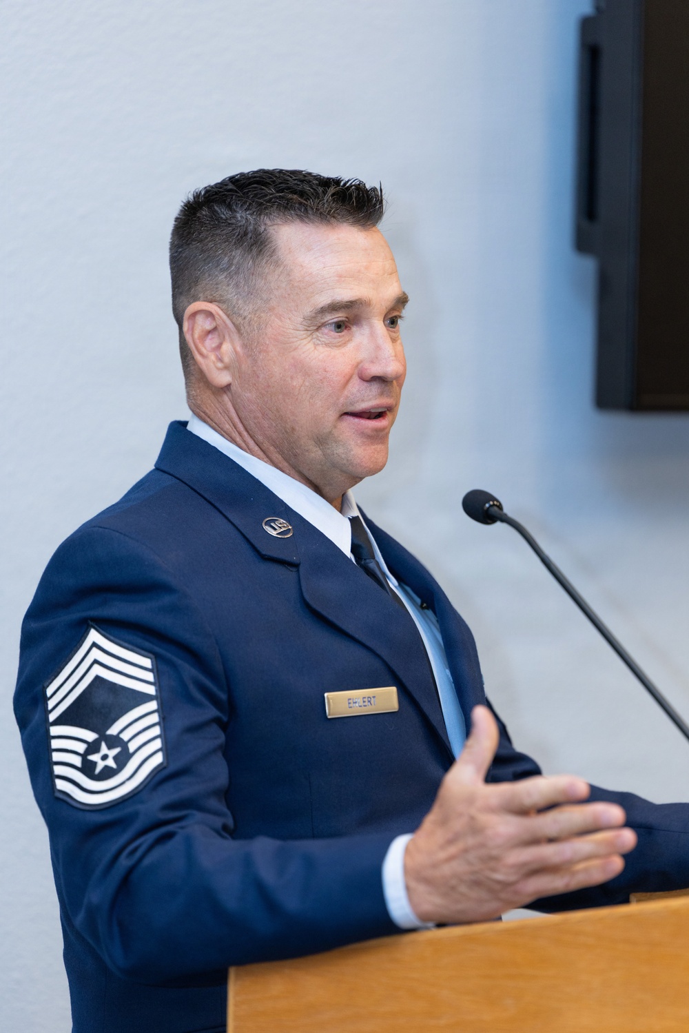 A U.S. Air Force Senior Master Sergeant named Ehlert is speaking at a podium, wearing the Air Force Service Dress uniform. He is gesturing with his right hand while addressing an audience, suggesting a formal ceremony or official presentation. His rank insignia and name tag are clearly visible on his uniform.