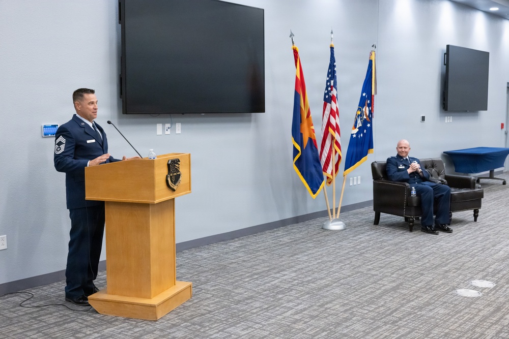 A military ceremony is taking place in a modern conference or ceremonial room. A senior non-commissioned officer in US Air Force dress blues is speaking at a wooden podium bearing a military insignia, while a seated officer in the background listens. Three flags — the Arizona state flag, the US flag, and what appears to be the Air National Guard flag — are displayed prominently.
