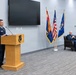 A military ceremony is taking place in a modern conference or ceremonial room. A senior non-commissioned officer in US Air Force dress blues is speaking at a wooden podium bearing a military insignia, while a seated officer in the background listens. Three flags — the Arizona state flag, the US flag, and what appears to be the Air National Guard flag — are displayed prominently.