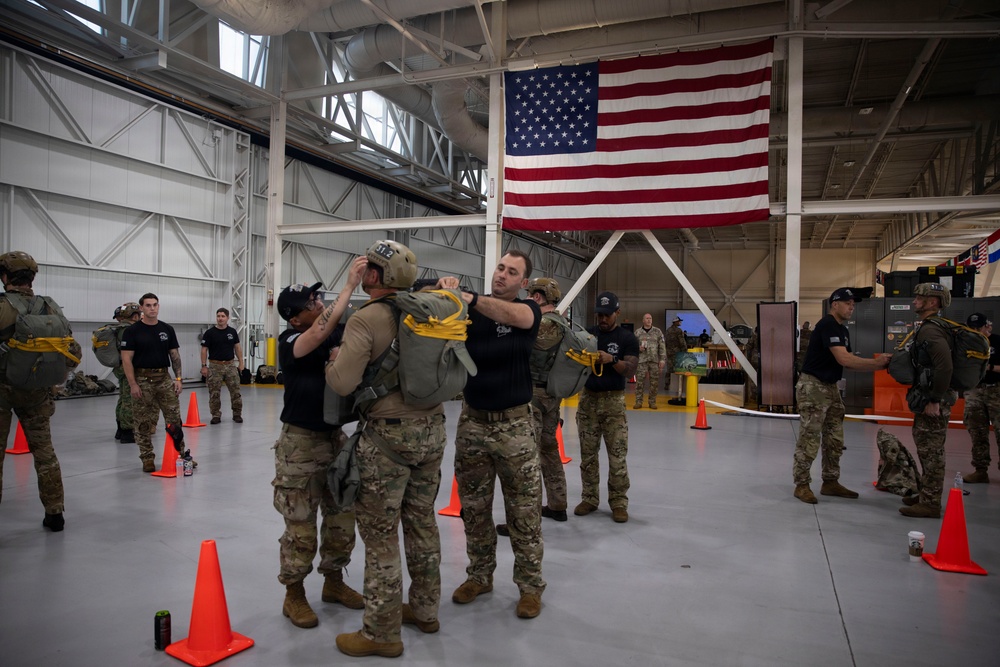 Military or law enforcement personnel are conducting equipment checks and parachute gear inspections inside a large hangar facility. A large American flag is prominently displayed on the rear wall of the hangar. Personnel wearing camouflage uniforms and tactical gear are organized in groups, with orange traffic cones marking designated areas on the floor.