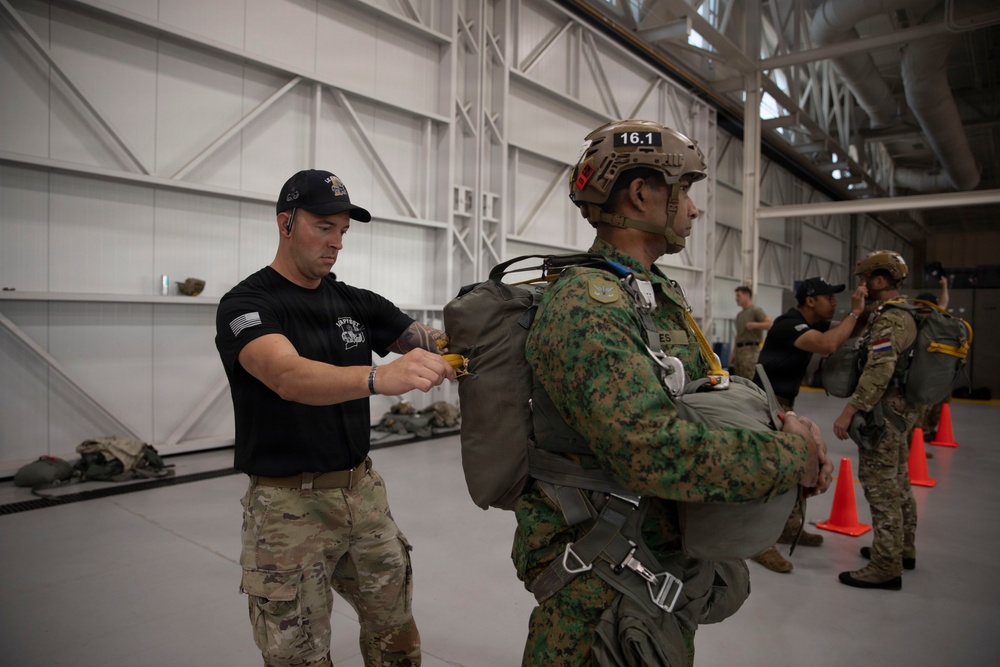 Military personnel are inside a large hangar facility conducting parachute equipment checks and inspections. A soldier in a black t-shirt with a US flag patch is inspecting or adjusting the parachute harness of another soldier wearing camouflage uniform and a tactical helmet marked '16.1'. Additional personnel and orange safety cones are visible in the background.