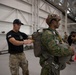 Military personnel are inside a large hangar facility conducting parachute equipment checks and inspections. A soldier in a black t-shirt with a US flag patch is inspecting or adjusting the parachute harness of another soldier wearing camouflage uniform and a tactical helmet marked '16.1'. Additional personnel and orange safety cones are visible in the background.