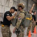 Military personnel are conducting parachute equipment checks inside a hangar facility. A soldier in a black t-shirt with a US flag patch is inspecting or adjusting the parachute harness of another soldier wearing Dutch (Netherlands) flag patch and a tactical helmet. Additional personnel in the background appear to be undergoing similar pre-jump equipment inspections.