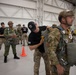 Military personnel are gathered inside a large hangar facility conducting parachute equipment checks and pre-jump preparations. A jumpmaster in a black cap and uniform is inspecting the parachute rig of a soldier wearing multicam uniform and a ballistic helmet. Multiple personnel in various military uniforms, including what appears to be a Singapore Armed Forces uniform, are present in the background.