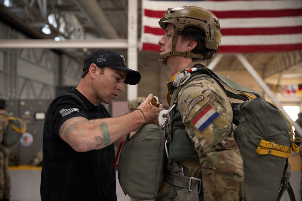 A military instructor or jumpmaster in civilian attire (black t-shirt and cap) is conducting a pre-jump equipment inspection on a soldier wearing full parachute gear and a tactical helmet. The soldier displays a French flag patch on their uniform sleeve, suggesting this is a multinational or allied training exercise. The setting appears to be a military hangar with a large American flag visible in the background.
