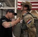 A military instructor or jumpmaster in civilian attire (black t-shirt and cap) is conducting a pre-jump equipment inspection on a soldier wearing full parachute gear and a tactical helmet. The soldier displays a French flag patch on their uniform sleeve, suggesting this is a multinational or allied training exercise. The setting appears to be a military hangar with a large American flag visible in the background.