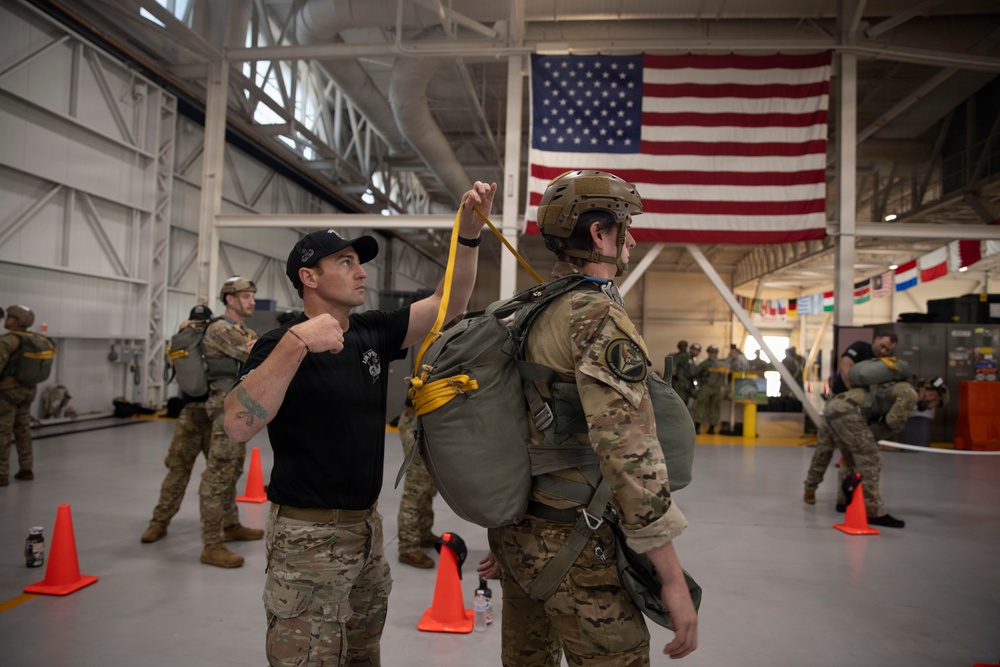 Military personnel are inside a large hangar facility conducting parachute equipment checks and inspections. A man in a black t-shirt and cap is inspecting or adjusting the yellow static line/ripcord of a soldier wearing full parachute gear, a tactical helmet, and combat uniform. A large American flag is prominently displayed in the background, along with international flags, suggesting a multinational military installation.
