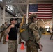 Military personnel are inside a large hangar facility conducting parachute equipment checks and inspections. A man in a black t-shirt and cap is inspecting or adjusting the yellow static line/ripcord of a soldier wearing full parachute gear, a tactical helmet, and combat uniform. A large American flag is prominently displayed in the background, along with international flags, suggesting a multinational military installation.