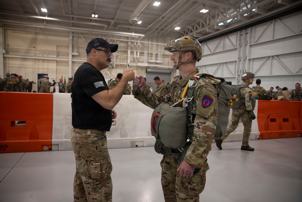 Two military personnel are fist-bumping inside a large hangar facility. One individual is in a black shirt with a baseball cap, while the other is in full multicam combat uniform with parachute equipment and a tactical helmet. The background shows numerous other military personnel in similar gear, suggesting a pre-jump or airborne operation staging event.