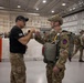 Two military personnel are fist-bumping inside a large hangar facility. One individual is in a black shirt with a baseball cap, while the other is in full multicam combat uniform with parachute equipment and a tactical helmet. The background shows numerous other military personnel in similar gear, suggesting a pre-jump or airborne operation staging event.