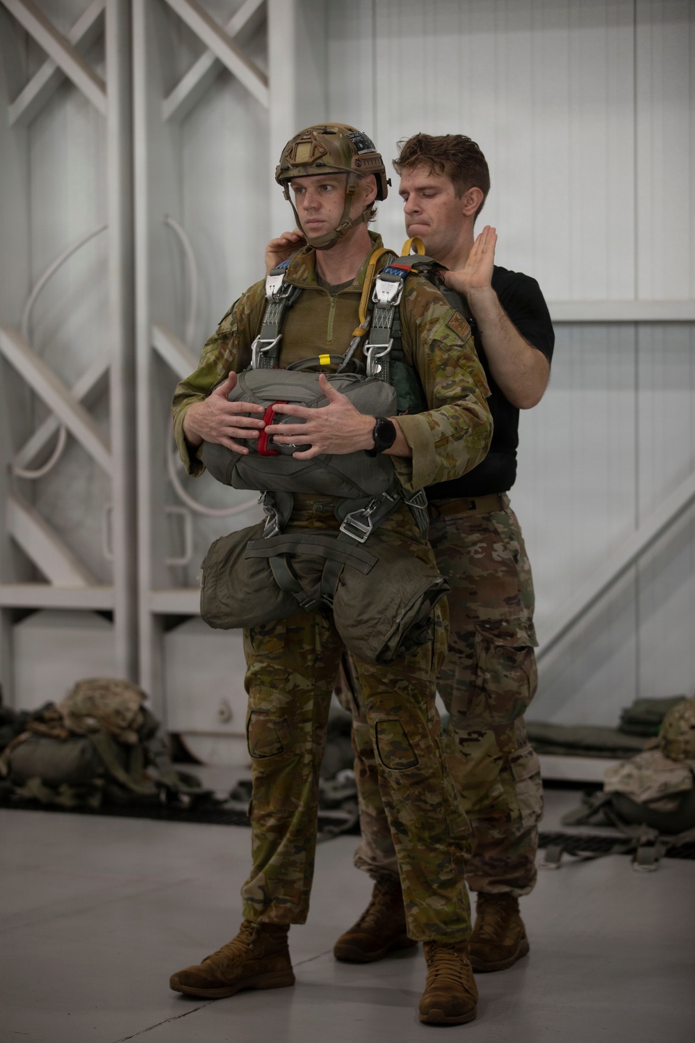 Two military personnel are shown in what appears to be an indoor training facility. One soldier wearing full parachute equipment, a tactical helmet, and multicam camouflage uniform is being inspected or fitted by a second individual in a black t-shirt and multicam trousers. The activity appears to be a pre-jump equipment check or parachute rigging inspection.