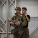 Two military personnel are shown in what appears to be an indoor training facility. One soldier wearing full parachute equipment, a tactical helmet, and multicam camouflage uniform is being inspected or fitted by a second individual in a black t-shirt and multicam trousers. The activity appears to be a pre-jump equipment check or parachute rigging inspection.