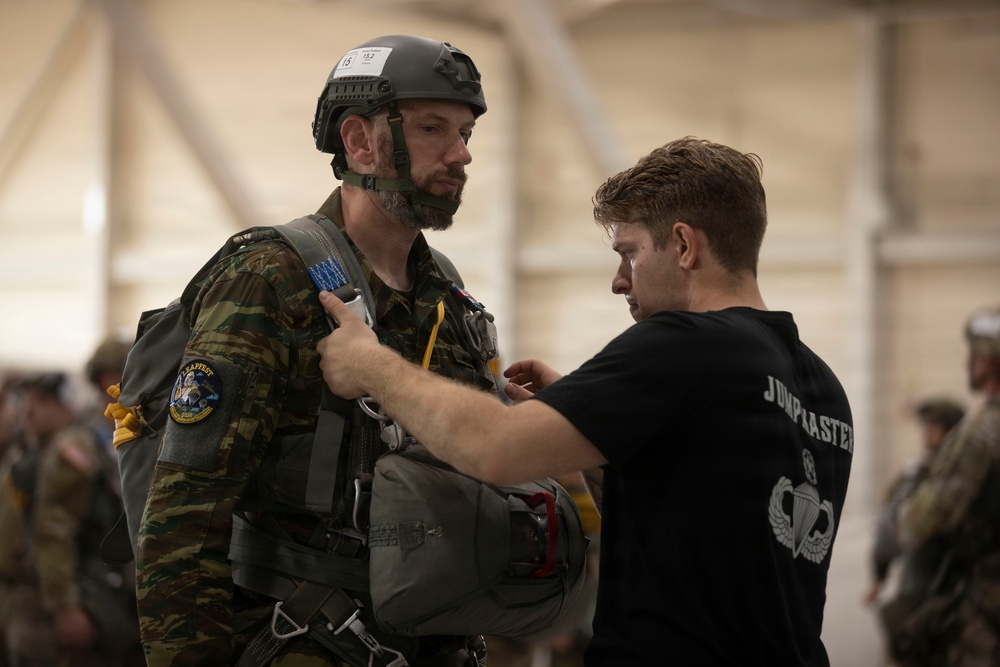 A military jumpmaster in a black t-shirt is conducting a pre-jump equipment inspection on a soldier wearing camouflage uniform, tactical helmet, and parachute harness. The inspection appears to be taking place inside a large hangar or staging area with other military personnel visible in the background. This is consistent with standard airborne operations pre-jump procedures.