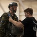 A military jumpmaster in a black t-shirt is conducting a pre-jump equipment inspection on a soldier wearing camouflage uniform, tactical helmet, and parachute harness. The inspection appears to be taking place inside a large hangar or staging area with other military personnel visible in the background. This is consistent with standard airborne operations pre-jump procedures.