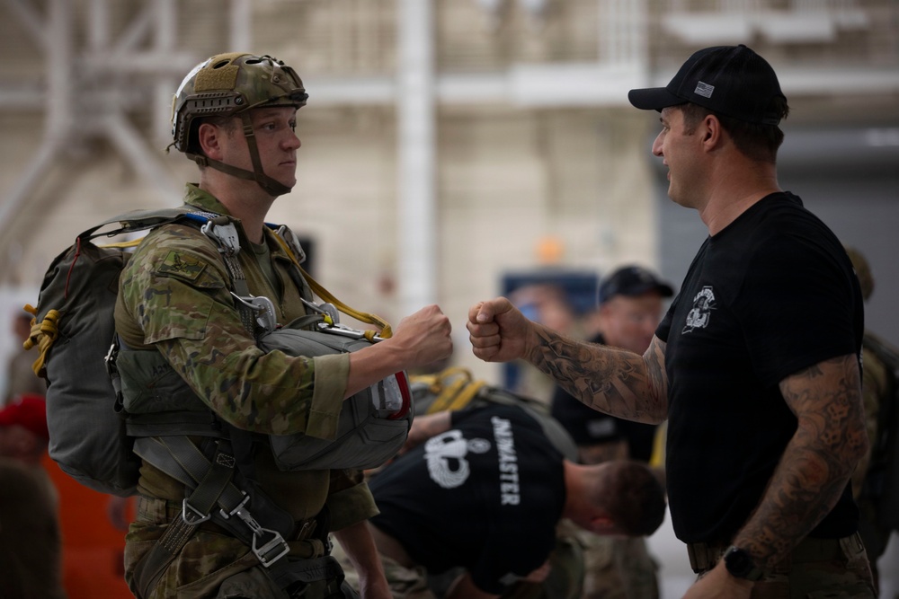 Two individuals are exchanging a fist bump in what appears to be a military hangar or staging area. The person on the left is wearing full combat/parachute gear including a tactical helmet, camouflage uniform, and a parachute harness with yellow straps. The person on the right is wearing a black t-shirt, black cap with a US flag patch, and has visible tattoos on both arms.