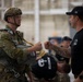 Two individuals are exchanging a fist bump in what appears to be a military hangar or staging area. The person on the left is wearing full combat/parachute gear including a tactical helmet, camouflage uniform, and a parachute harness with yellow straps. The person on the right is wearing a black t-shirt, black cap with a US flag patch, and has visible tattoos on both arms.