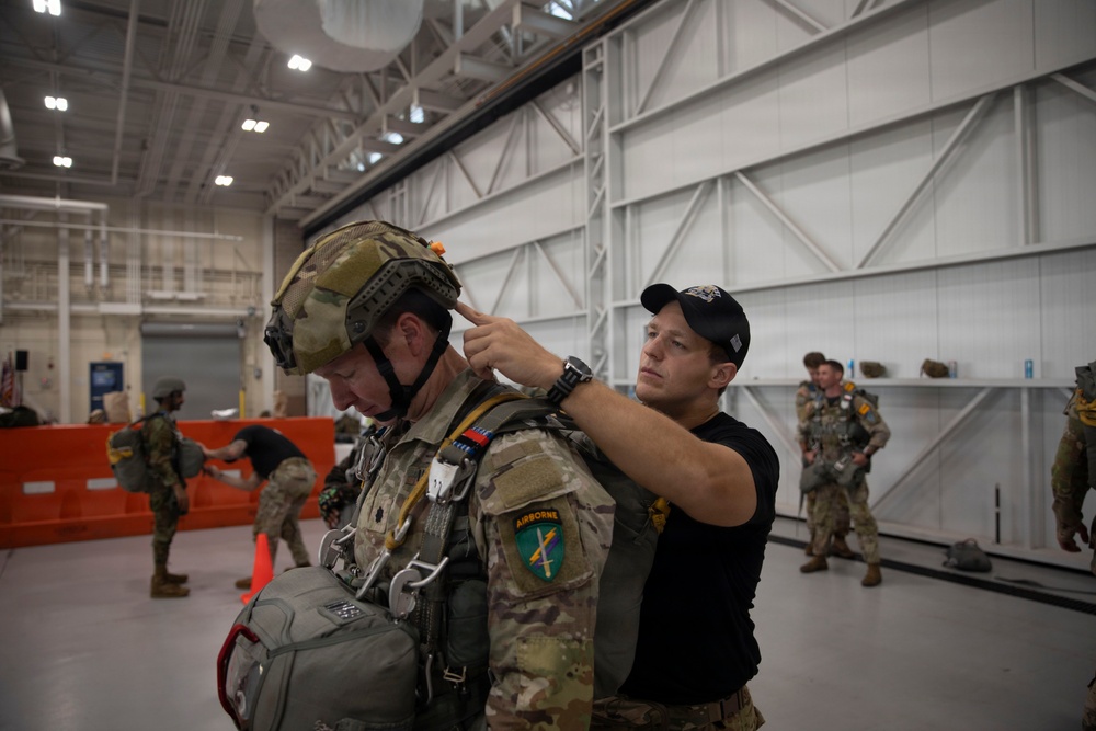 A military personnel in full combat gear including a tactical helmet and parachute harness is being inspected or assisted by a man in civilian-style black clothing and cap inside what appears to be a military hangar. The soldier wears an Airborne patch and multicam/OCP uniform with full jump equipment. Additional soldiers in similar gear are visible in the background of the large industrial facility.