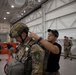 A military personnel in full combat gear including a tactical helmet and parachute harness is being inspected or assisted by a man in civilian-style black clothing and cap inside what appears to be a military hangar. The soldier wears an Airborne patch and multicam/OCP uniform with full jump equipment. Additional soldiers in similar gear are visible in the background of the large industrial facility.
