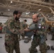 Two military personnel in camouflage uniforms are inside a large hangar, assisting each other with parachute or jump equipment preparation. One individual displays a Qatar flag patch on their uniform sleeve. Multiple other soldiers are visible in the background engaged in similar equipment checks, suggesting a multinational airborne training exercise.