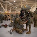 Military personnel in camouflage uniforms are preparing parachute equipment inside a large hangar facility. Multiple soldiers are assisting each other with gear checks and harness adjustments prior to what appears to be a parachute operation. The presence of multiple national flags and varied uniform patterns suggests this is a multinational military exercise.