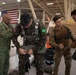 Military personnel from multiple nations are inside a hangar conducting parachute equipment checks and rigging. A soldier wearing a Brazilian flag patch is being assisted with his parachute harness by a colleague bearing an 82nd Airborne Division patch. The setting appears to be a joint multinational airborne training exercise, evidenced by multiple national flags visible in the background.