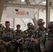 A group of military paratroopers in full jump gear, including parachute packs and helmets, are gathered inside a hangar with heads bowed, apparently in a pre-jump prayer or moment of reflection. A banner in the background reads 'LEAPFEST,' which is an international military parachuting competition hosted annually by the U.S. Army. The personnel are wearing various camouflage uniforms suggesting participation from multiple nations.