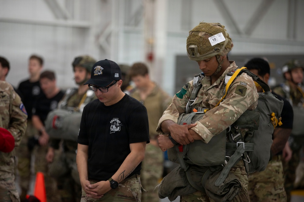 The image shows a military setting, likely a hangar, where personnel in various uniforms are gathered. A fully equipped paratrooper wearing desert camouflage, a tactical helmet with a numbered tag (10), and a parachute harness stands prominently in the foreground alongside a civilian or lightly dressed individual in a black cap and t-shirt. The scene appears to be a pre-jump inspection or briefing event involving multinational military personnel.