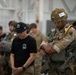 The image shows a military setting, likely a hangar, where personnel in various uniforms are gathered. A fully equipped paratrooper wearing desert camouflage, a tactical helmet with a numbered tag (10), and a parachute harness stands prominently in the foreground alongside a civilian or lightly dressed individual in a black cap and t-shirt. The scene appears to be a pre-jump inspection or briefing event involving multinational military personnel.